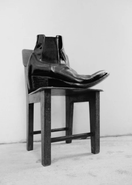 Black leather boot placed as the primary subject on a vintage-style wooden chair in a minimalist indoor setting against a white wall.