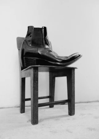 Black leather boot placed as the primary subject on a vintage-style wooden chair in a minimalist indoor setting against a white wall.