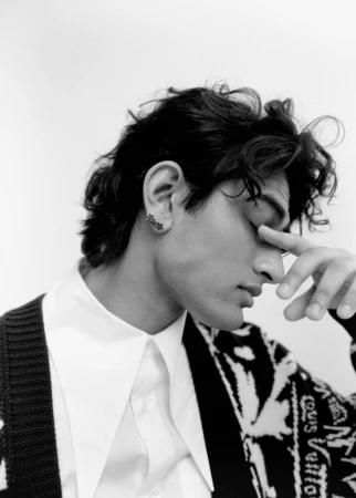 Close-up profile portrait of a man wearing a white collared shirt and patterned cardigan, touching his nose against a plain white background.