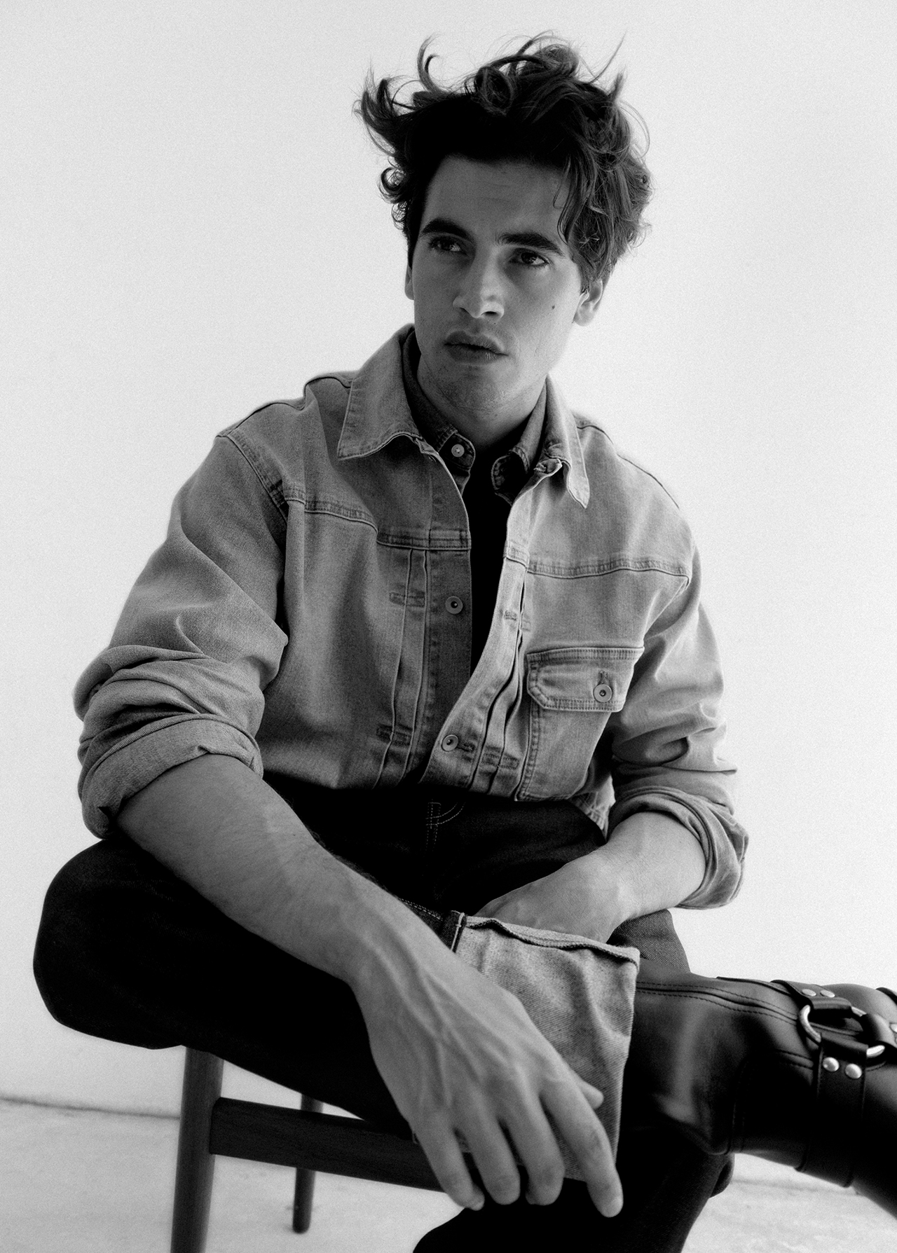 Monochrome portrait of a young man wearing a denim jacket and dark boots seated on a chair against a plain background