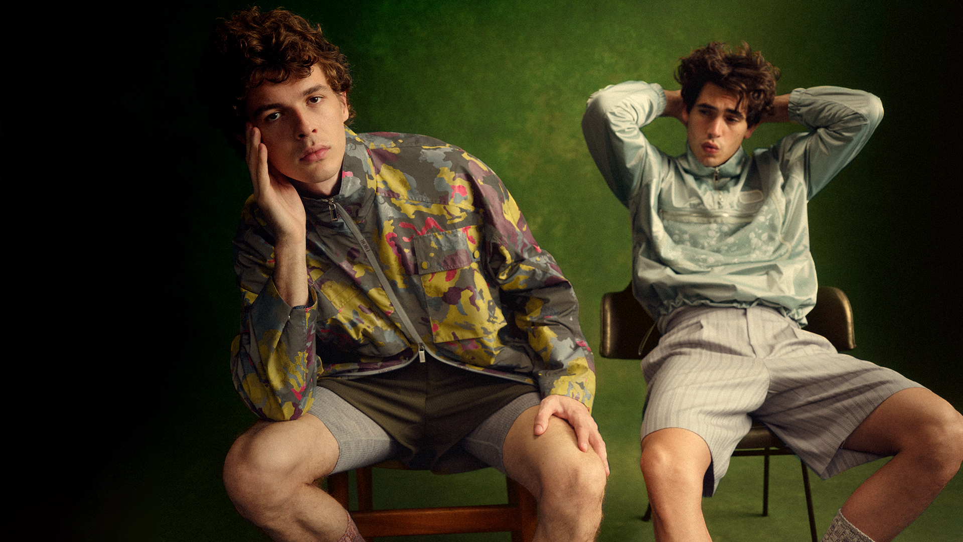 Two young men in patterned tops pose indoors against a mottled green backdrop, seated in chairs with a relaxed, casual vibe.