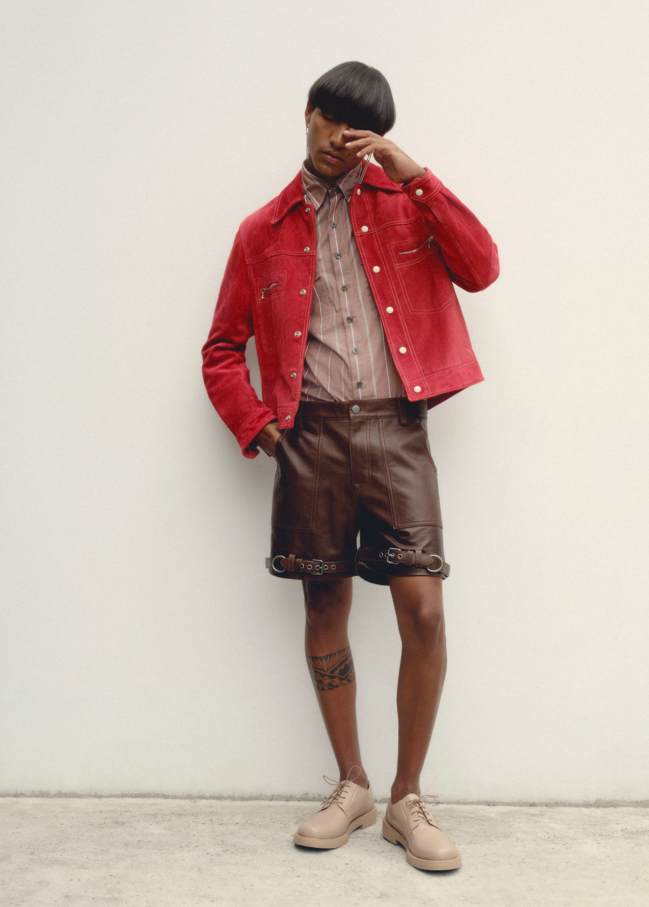 A person with a stylish haircut wearing a red jacket, striped shirt, and leather shorts poses against a simple white wall in soft lighting.