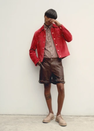 A person with a stylish haircut wearing a red jacket, striped shirt, and leather shorts poses against a simple white wall in soft lighting.