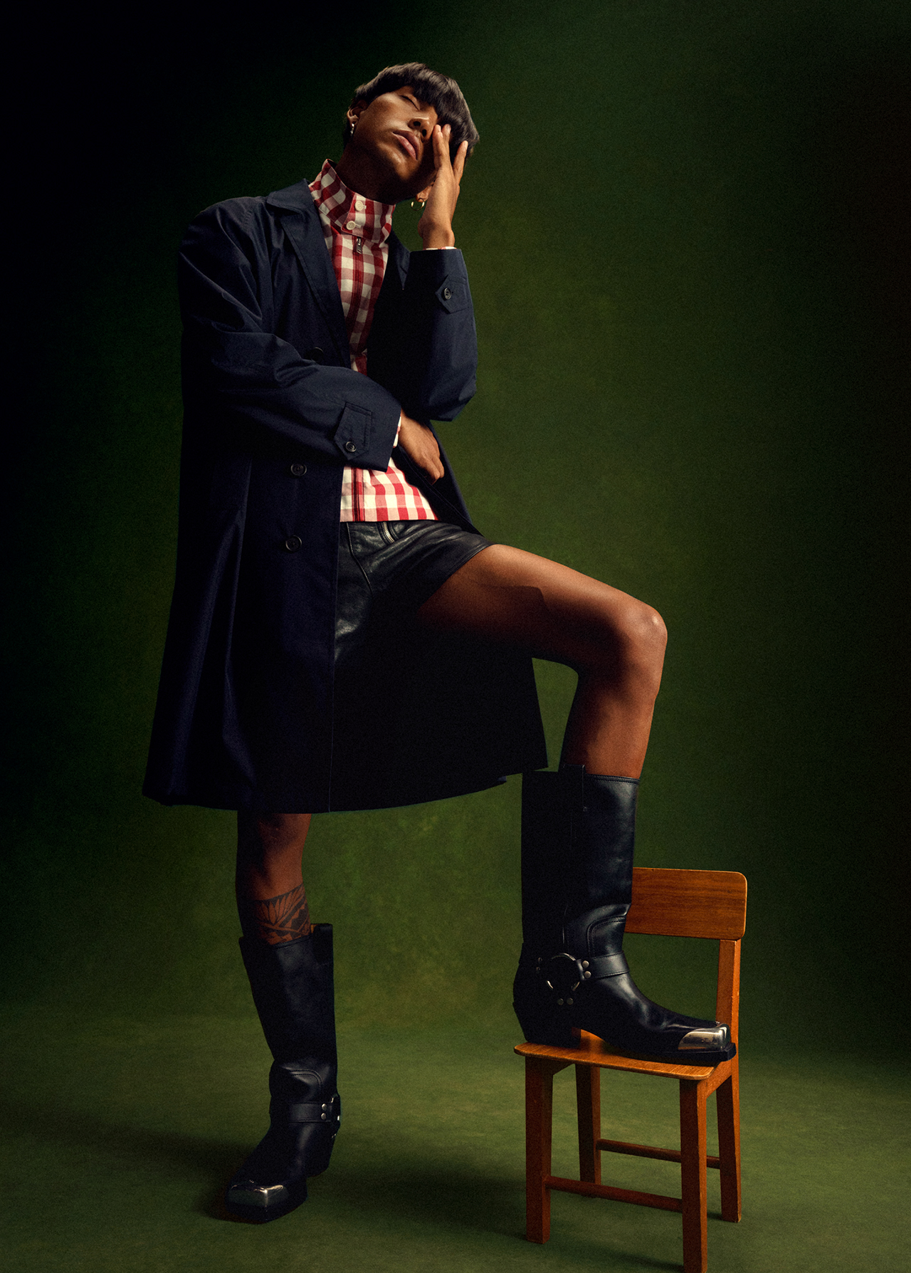 A person wearing a coat, plaid shirt, shorts, and boots sits on a wooden chair against a green backdrop in a fashion pose.