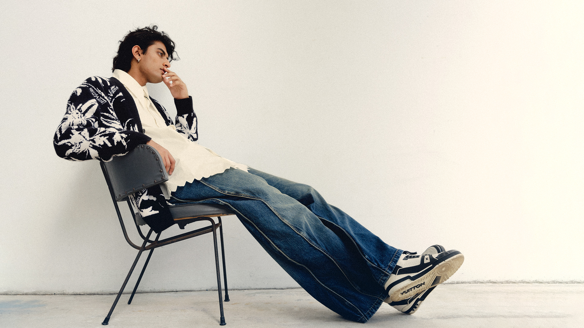 A young adult male with dark hair sits in profile on a gray chair in a cream-colored room, wearing jeans, patterned cardigan, and sneakers.