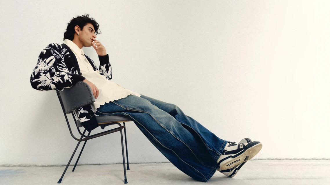 A young adult male with dark hair sits in profile on a gray chair in a cream-colored room, wearing jeans, patterned cardigan, and sneakers.