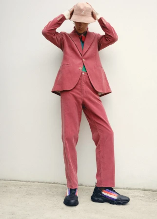 Person in a corduroy suit and bucket hat poses against a white wall, standing in black sneakers with colorful stripes, full shot.