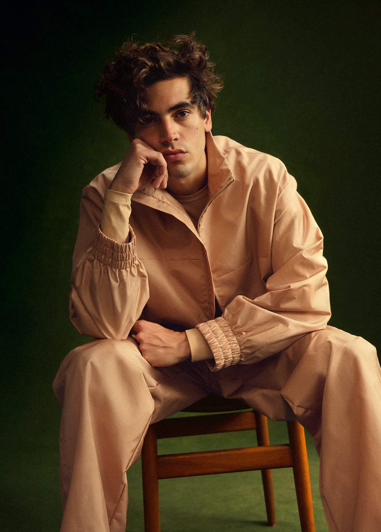 Young adult male with dark, curly hair, wearing a pink jacket and pants, posing on a wooden chair against a dark green background.
