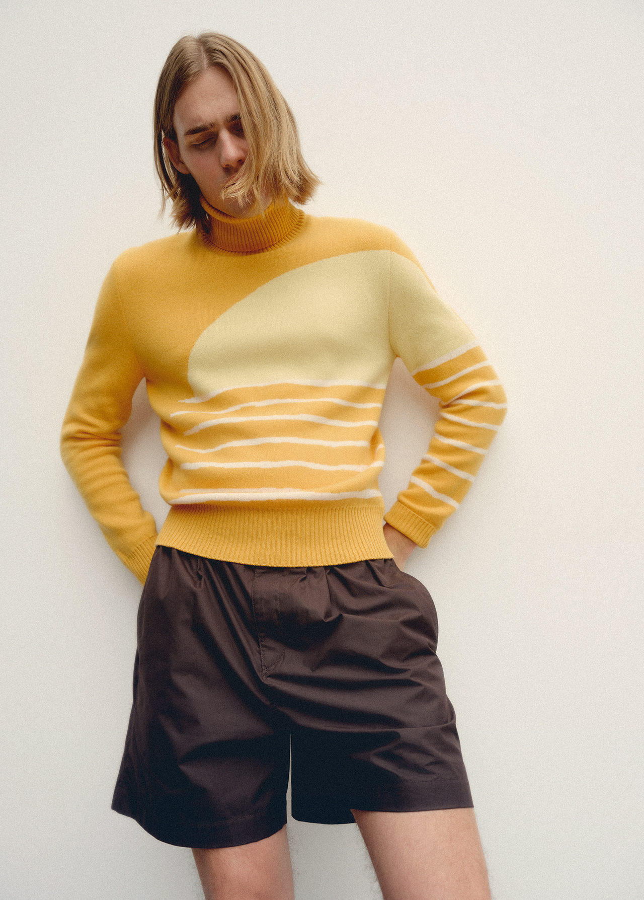 Male fashion model wears a yellow and cream sweater and dark brown shorts in a studio shot against a light neutral background.