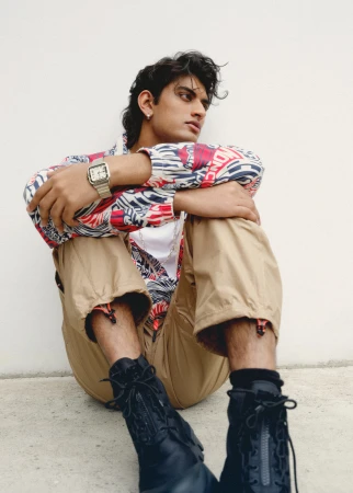Young man sitting on the ground wearing patterned jacket, tan trousers and black boots against a plain wall