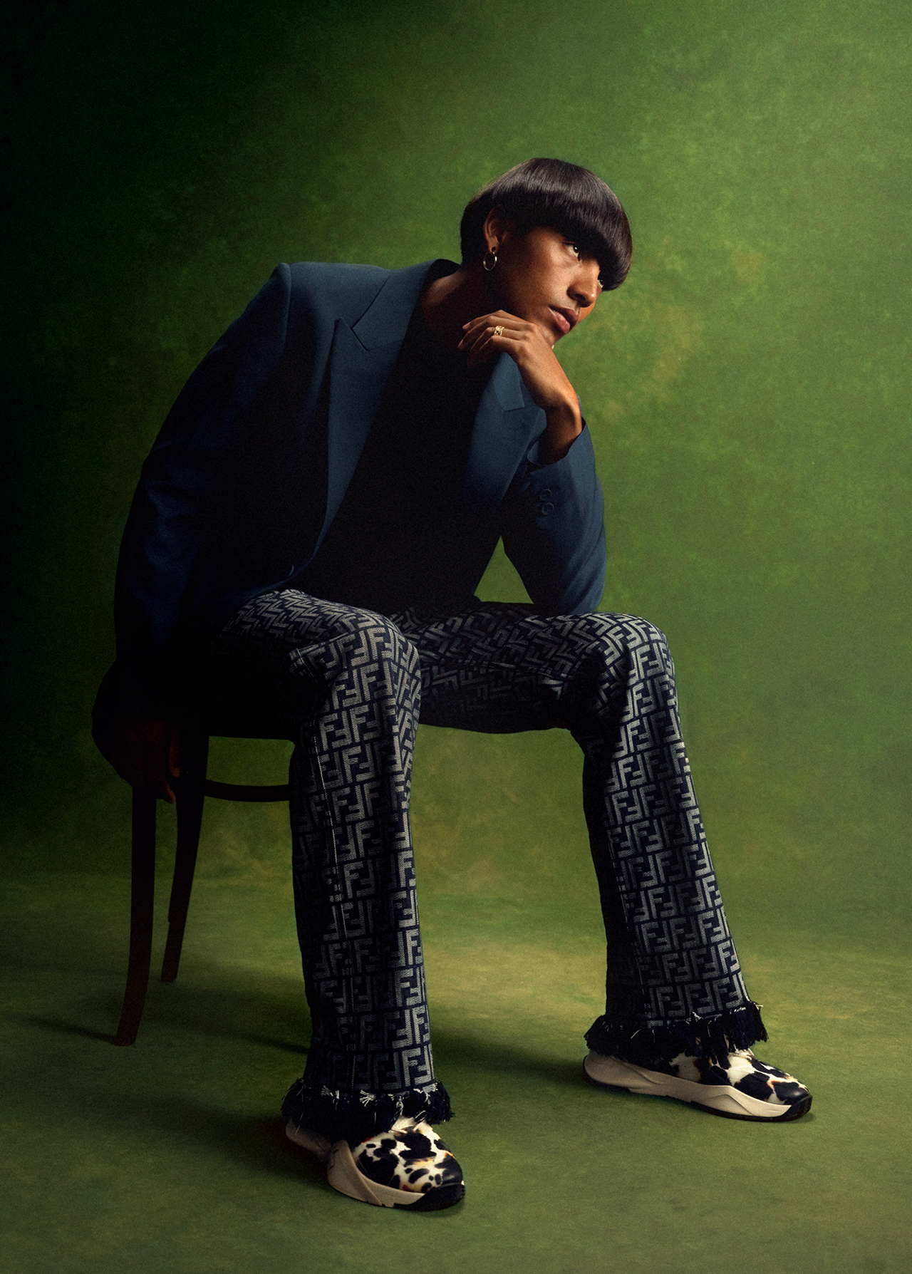 A person sits in a chair wearing patterned pants and a blue blazer in a portrait against an olive green backdrop in a studio.