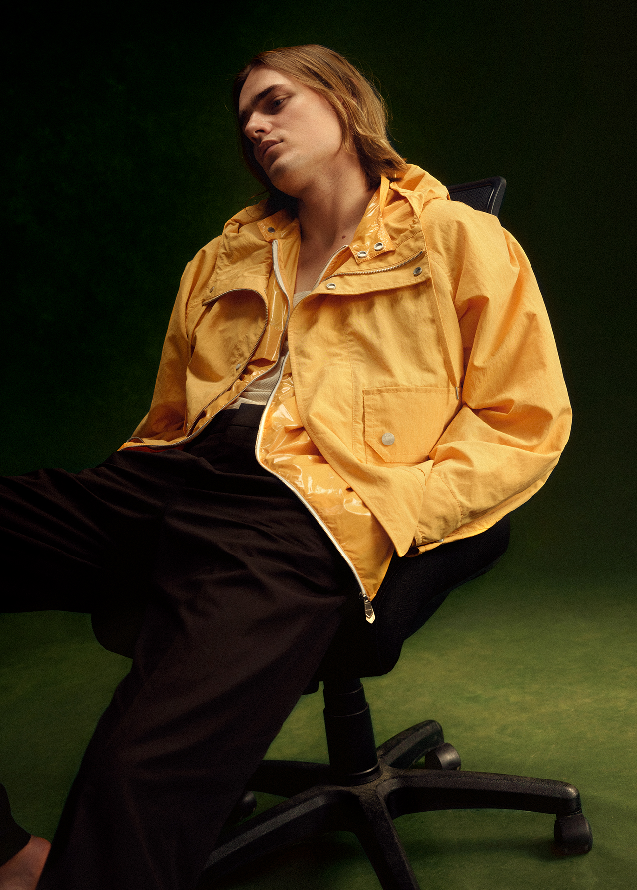 Model wearing a yellow jacket and dark trousers seated on a black office chair against a textured dark green background