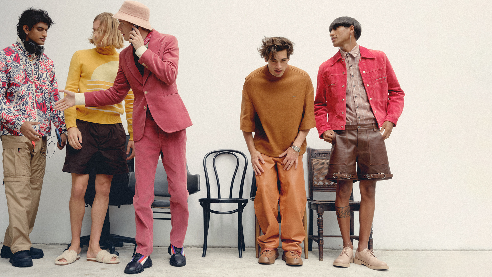 Five stylish men in fashionable outfits pose indoors against a white wall, showcasing unique textures and color palettes in a contemporary fashion shoot.