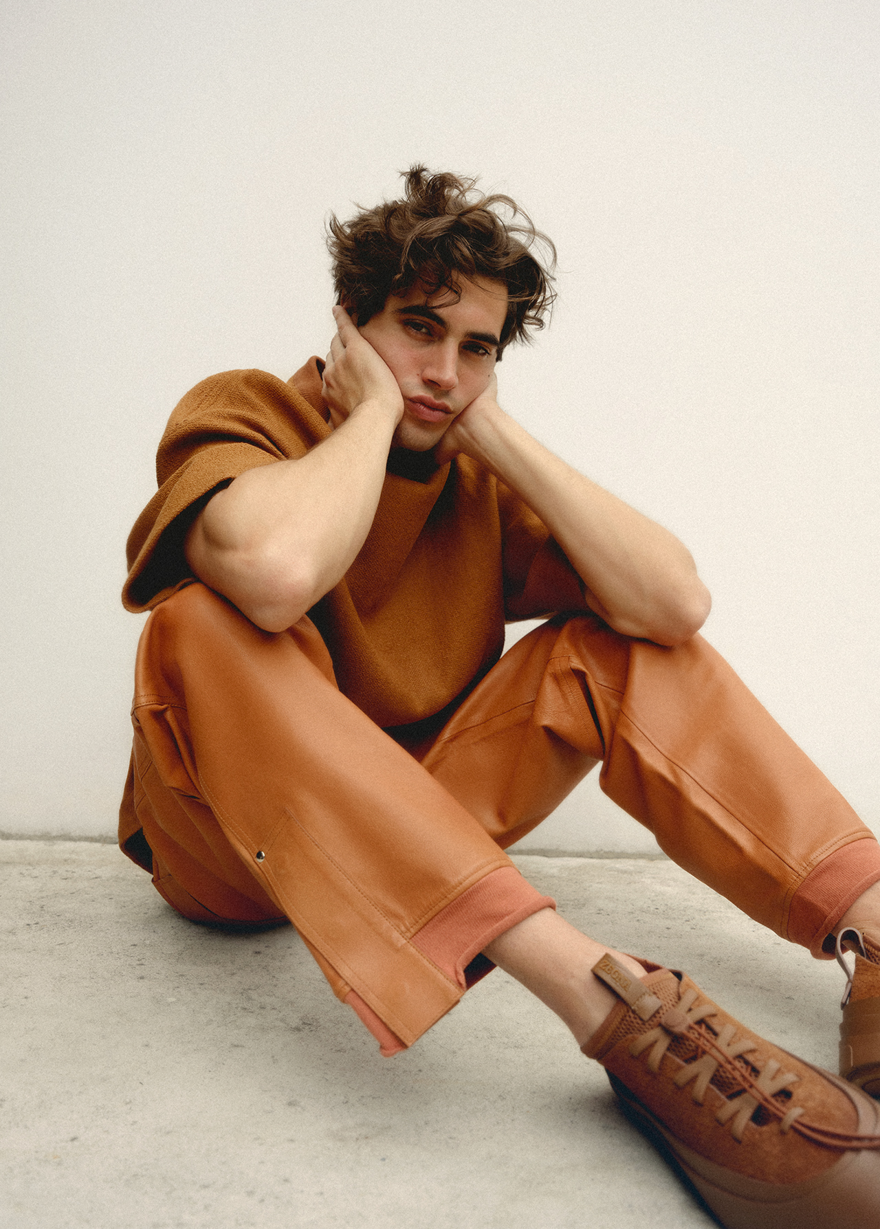 A young adult male with dark hair sits indoors, wearing a brown shirt and pants, with brown shoes, his hands resting on his face.