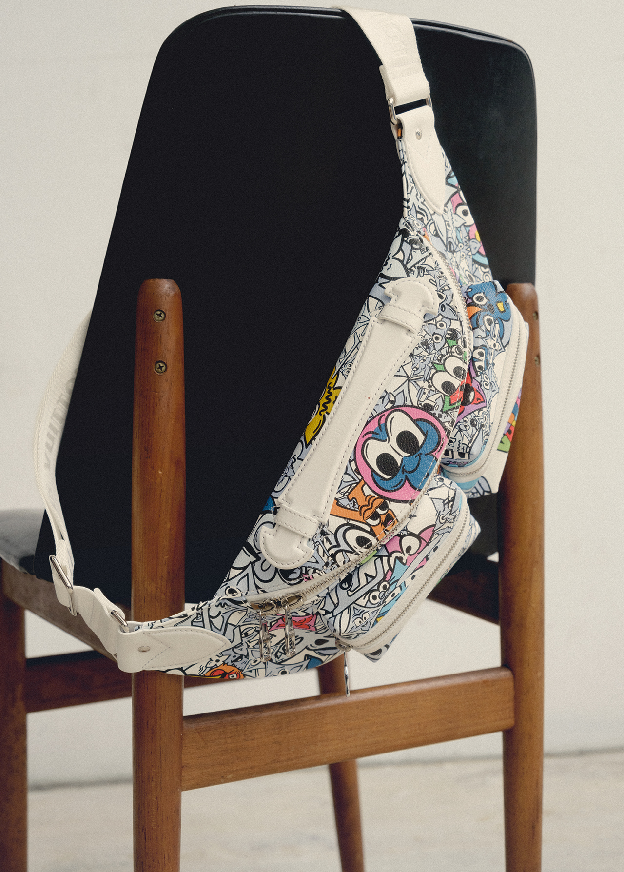 Designer handbag with doodle-style cartoon graphics on a wooden chair against a plain backdrop in a studio setting.