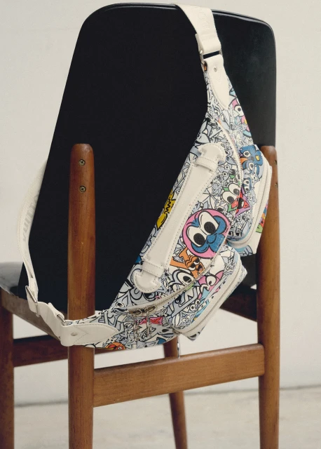 Designer handbag with doodle-style cartoon graphics on a wooden chair against a plain backdrop in a studio setting.