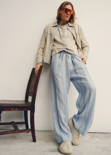Man in a casual outfit featuring a linen jacket, t-shirt, and striped trousers, standing indoors near a chair in a fashion pose.