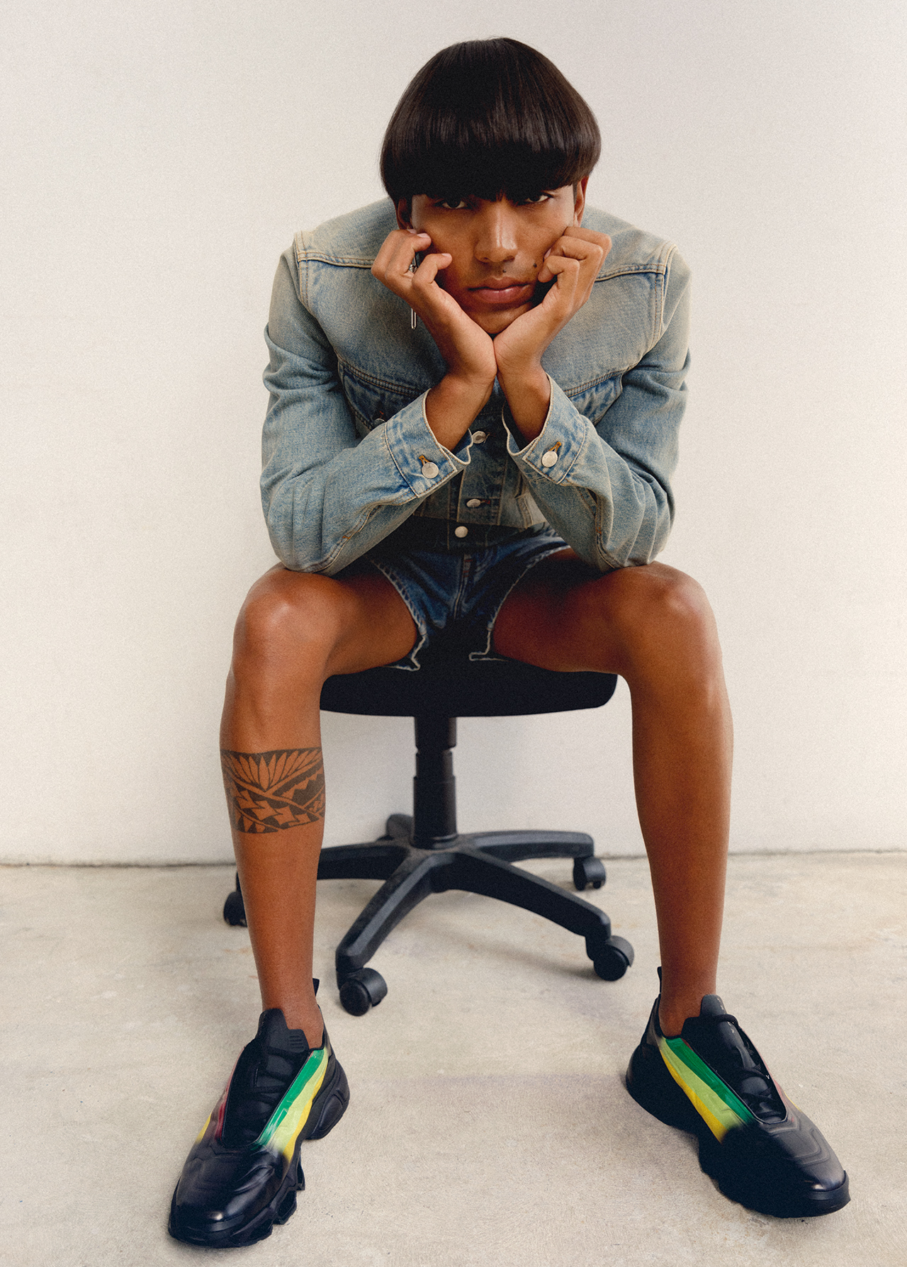 A young adult sits on an office chair, hands supporting their face, wearing a denim jacket and shorts with black sneakers in a studio.