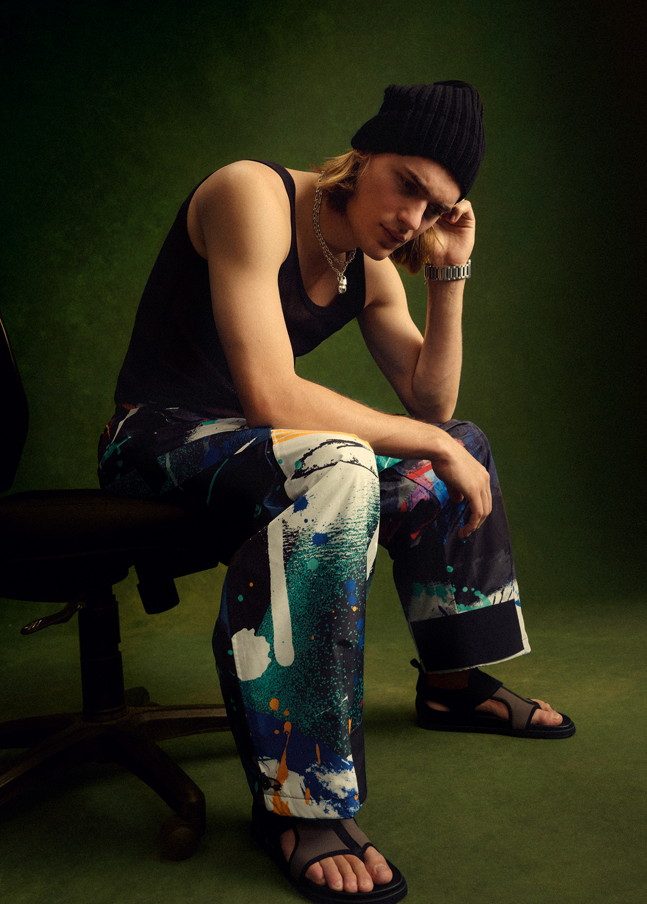 A young adult male sits indoors on a chair wearing a black tank top, colorfully splattered pants, sandals, and a black beanie.