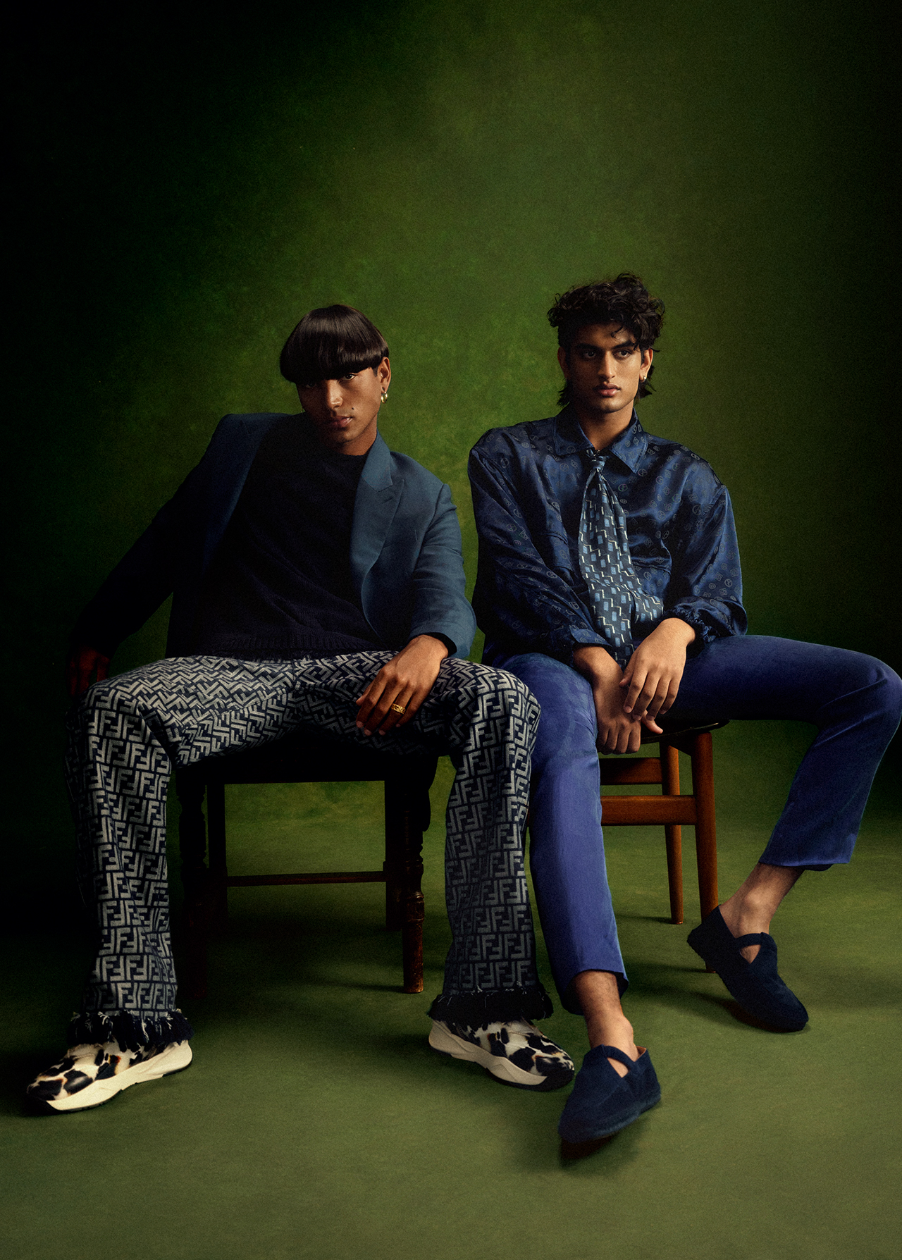 Two stylish men in patterned suits pose indoors against a green backdrop, seated on wooden chairs, showcasing unique hairstyles and fashion.