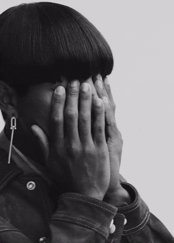 Close-up portrait of a non-binary person with a bowl haircut covering their eyes with their hands in a black and white studio setting.