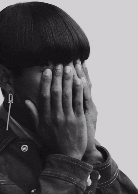 Close-up portrait of a non-binary person with a bowl haircut covering their eyes with their hands in a black and white studio setting.