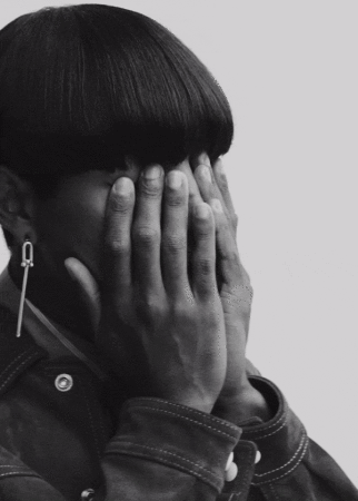 Close-up portrait of a non-binary person with a bowl haircut covering their eyes with their hands in a black and white studio setting.