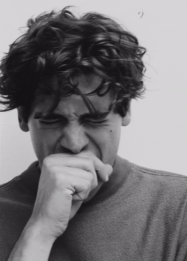 Monochrome portrait of a young adult yawning with a hand covering their mouth, set against a neutral background in an indoor setting.