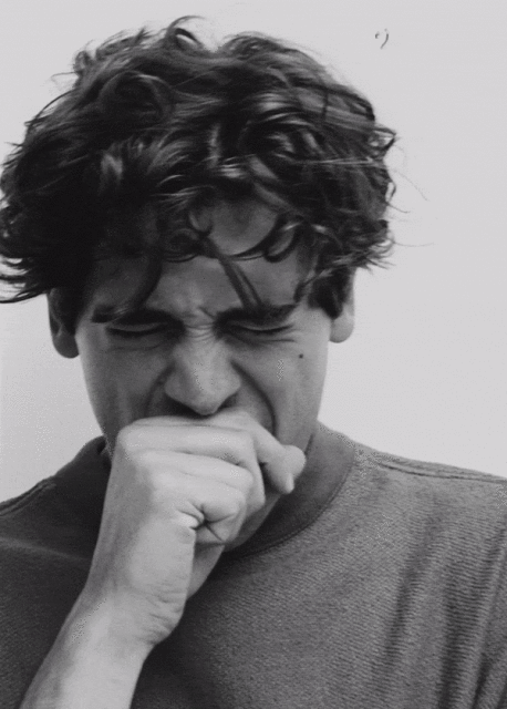 Monochrome portrait of a young adult yawning with a hand covering their mouth, set against a neutral background in an indoor setting.