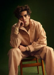A man with curly hair seated on a wooden chair wearing a beige zip-up jacket and matching trousers.