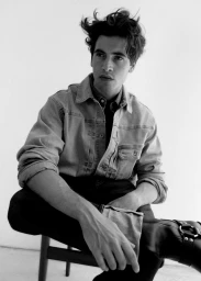 Monochrome portrait of a young man wearing a denim jacket and dark boots seated on a chair against a plain background