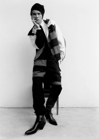 Monochrome portrait of a man in a beanie and patterned jacket leaning against a chair against a white background