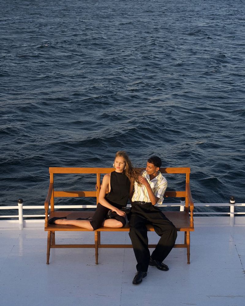 Woman and man in stylish outfits are sitting on a wooden bench on the deck of a boat in the sea with deep blue water.