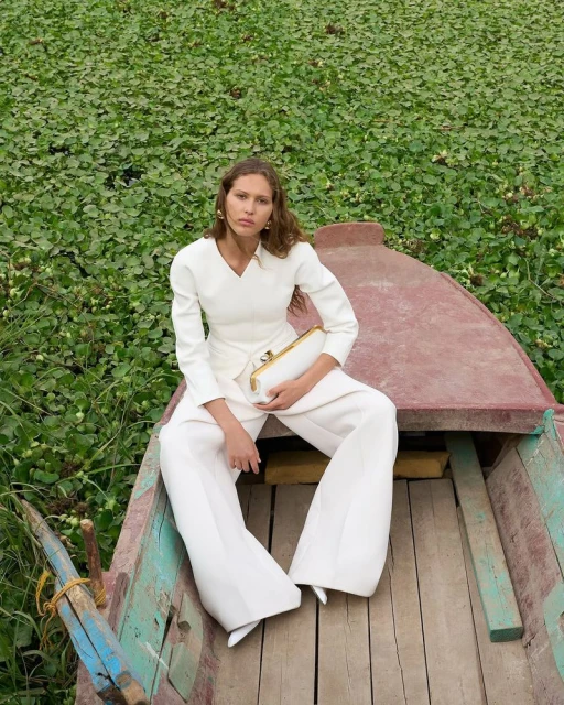 An adult female model sits in a wooden boat with white clothing and purse, amid green aquatic plants. The image highlights fashion in a natural setting.