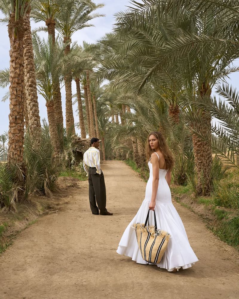 A woman in a white dress and a man are walking on a path lined with palm trees. The woman is carrying a beach bag.