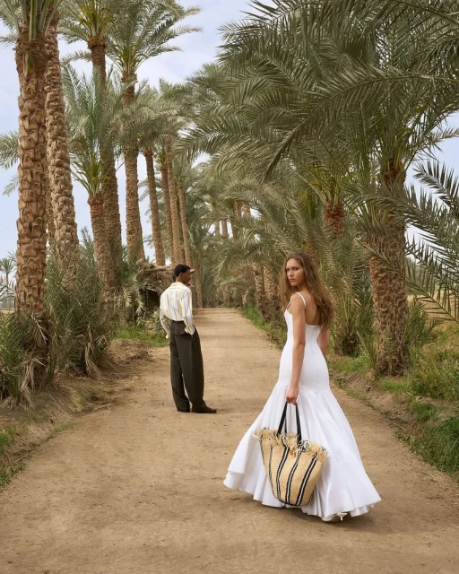 A woman in a white dress and a man are walking on a path lined with palm trees. The woman is carrying a beach bag.