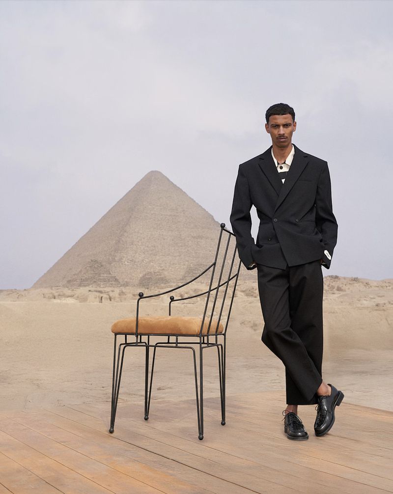 Fashion shot of a man in a black suit standing next to a chair in the desert with a pyramid in the background on a cloudy day.