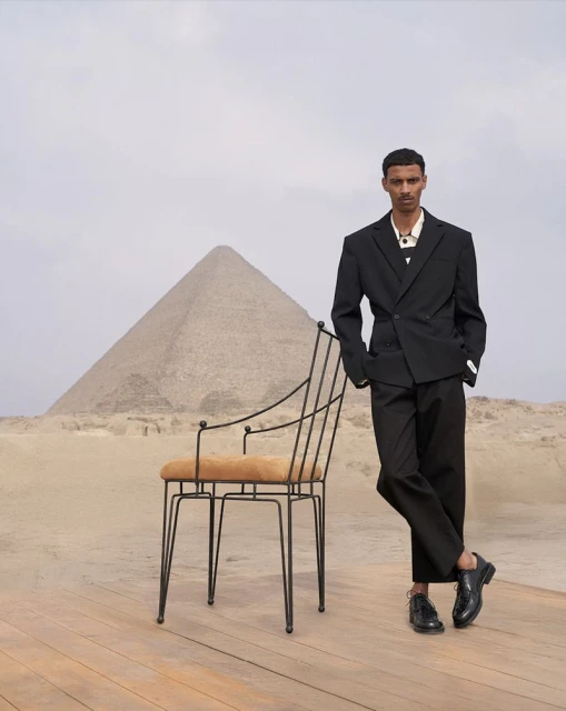 Fashion shot of a man in a black suit standing next to a chair in the desert with a pyramid in the background on a cloudy day.