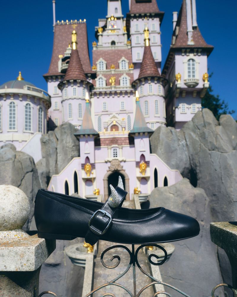 Black Mary Jane style shoe with bow, positioned on a stone ledge with a Disney-style castle in the background on a sunny day.