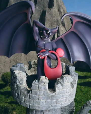 A statue of gargoyle-like creature standing on a castle tower with a red handbag in front of a mountain landscape.