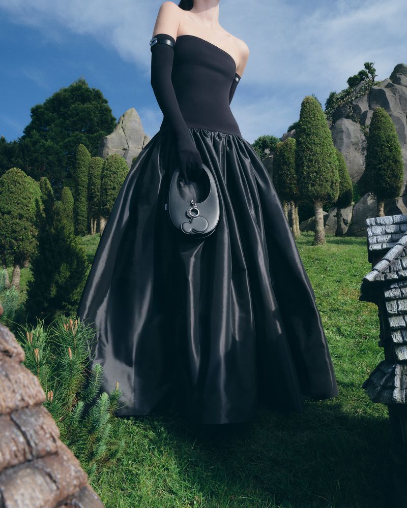 Woman in elegant black dress and gloves posing outdoors with handbag, set against green grass and trees under a blue sky. Fashion editorial.