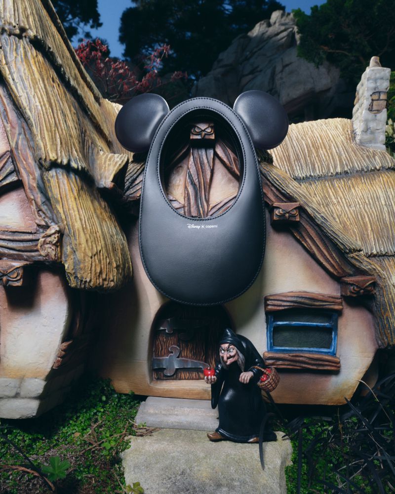 Whimsical still life of a black Mickey Mouse-shaped handbag in front of a storybook house, with a Disney villain figurine beside the entrance.