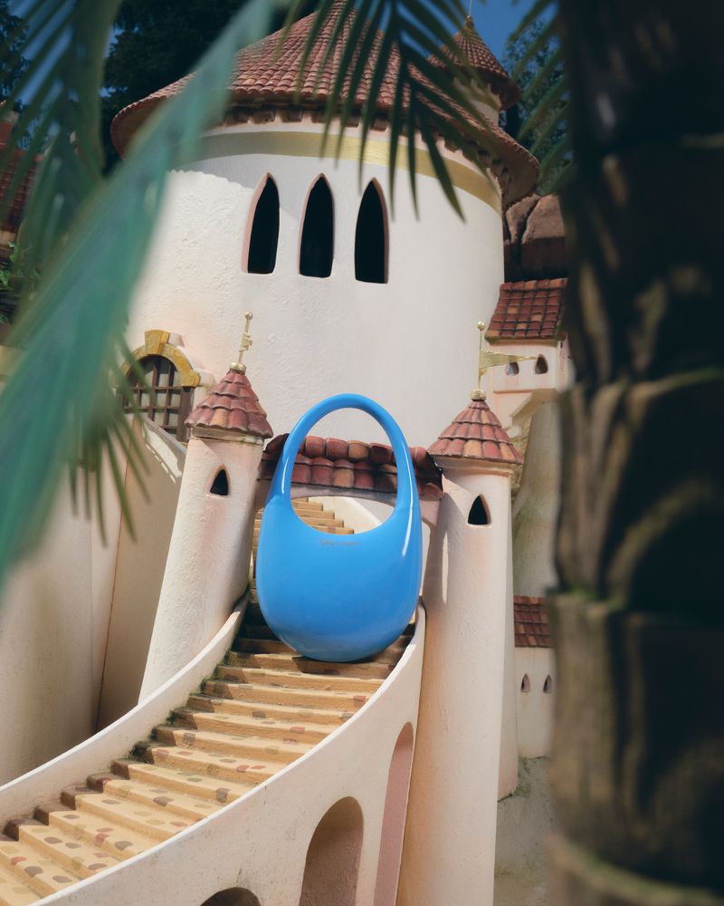 Blue purse sits on tan stone stairs of a sandcastle-like building with a red tile roof, viewed through green leaves.