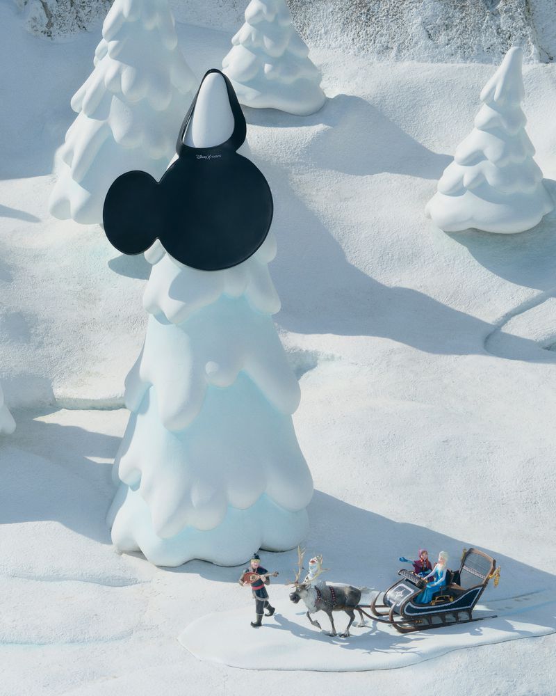 Winter holiday scene with a Minnie Mouse handbag on a snow-covered tree, a reindeer pulling a sleigh, and a figure walking in the snow.