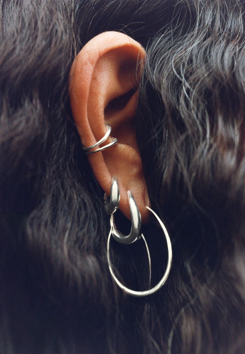 Close-up of an ear with multiple silver-colored earrings. Focus is on the jewelry and ear anatomy against dark wavy hair.