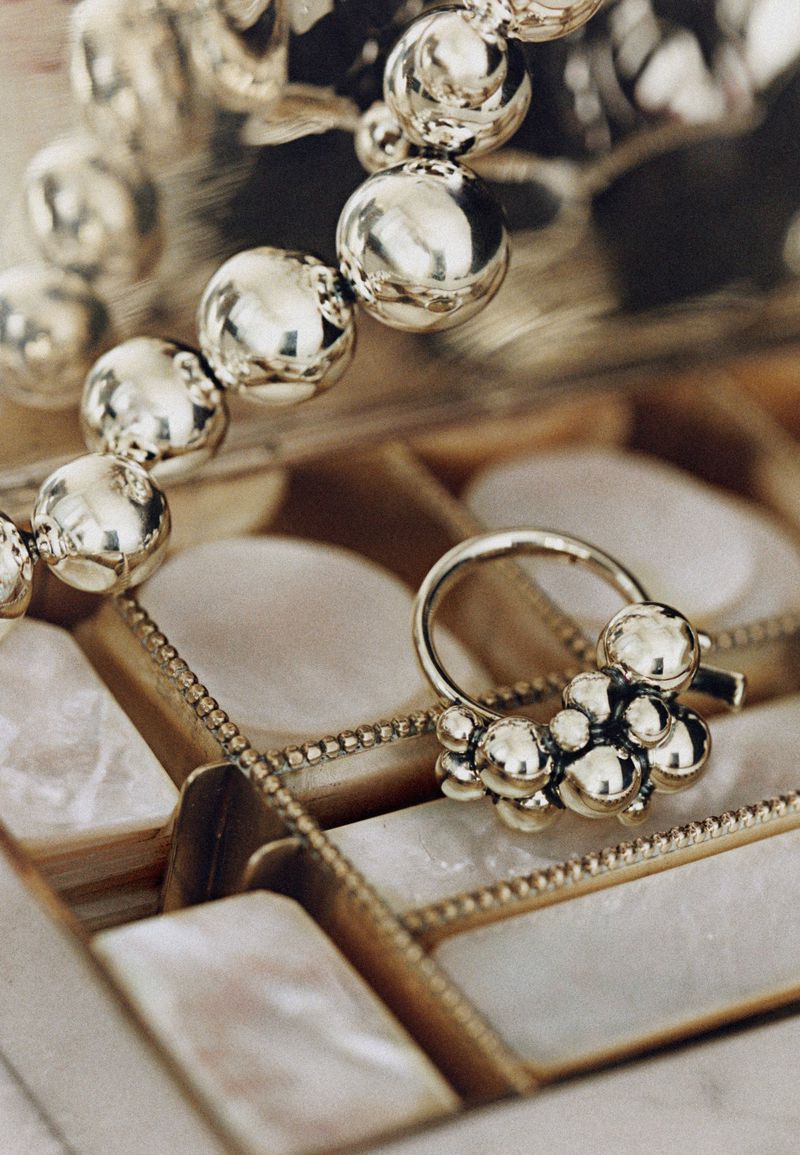 Close-up of an ornate silver jewelry set with a ring and beaded necklace displayed inside of a partitioned jewelry box with pearl accents.