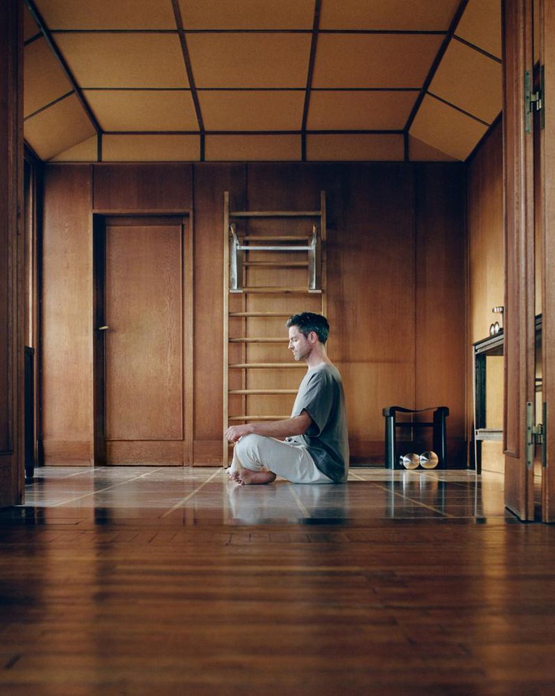 Adult male meditates cross-legged on wood floor in wood-paneled room with ladder. Man wears a gray shirt and white pants.