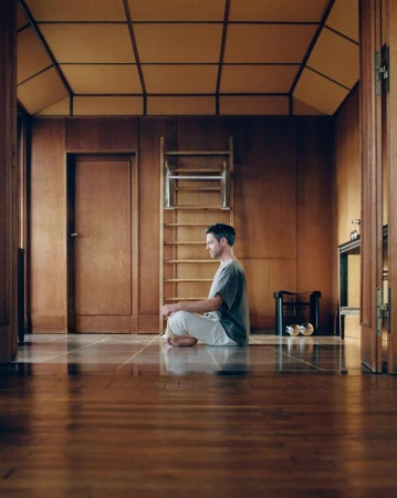 Adult male meditates cross-legged on wood floor in wood-paneled room with ladder. Man wears a gray shirt and white pants.