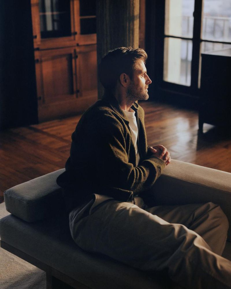 Man sitting on a couch in a sunlit room, wearing a cardigan and tan pants, looking towards the window with natural light and hardwood floors.