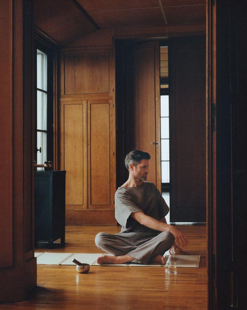 Adult man in grey loungewear sits cross-legged on a mat inside a wood-paneled room, meditating with a bowl nearby in a tranquil setting.